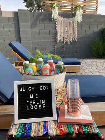 Outdoor poolside patio with blue lounge chairs, basket of colorful bottled juices on ice, stacked plastic cups and napkins, a black letter board reading 'JUICE GOT ME FEELIN LOOSE', and a boho macramé wall hanging on a cinderblock fence.