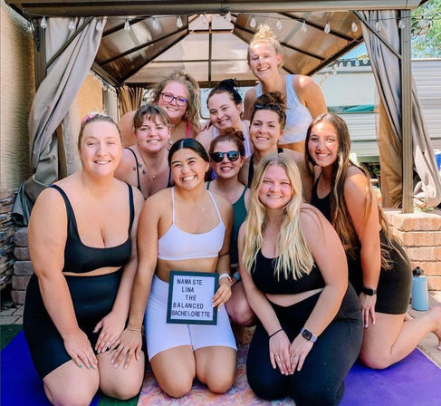 Smiling group of women in activewear at an outdoor backyard gazebo bachelorette yoga party, posing on yoga mats and holding a small letter board with a playful 'namaste' bachelorette message.
