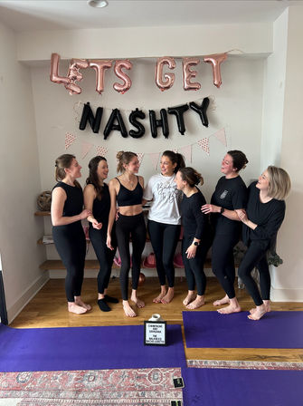 Seven women in black activewear laughing together at an indoor bachelorette yoga party under rose-gold and black letter balloons reading “LET’S GET NASH TY,” standing on purple yoga mats on a wooden floor.