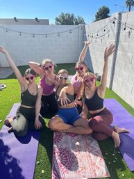 Group of friends in activewear posing on colorful yoga mats in a sunny backyard with string lights, wearing heart-shaped sunglasses and raising their arms for a playful outdoor yoga session on artificial grass