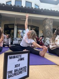 Outdoor bachelorette yoga class on a backyard patio — women stretching on purple mats with a playful lightbox bachelorette sign, stone columns and arched windows in the background.