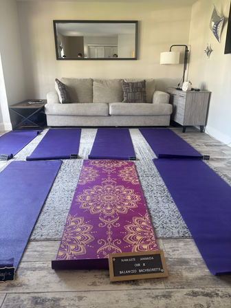 Cozy living room yoga setup with a light-gray sofa and wall mirror, six purple yoga mats laid out on the floor and a central purple-and-gold patterned mat with a small letterboard announcing a bachelorette yoga session; floor lamp, side tables and silver balloons in the background.