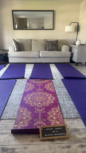 Cozy living room yoga setup with a light-gray sofa and wall mirror, six purple yoga mats laid out on the floor and a central purple-and-gold patterned mat with a small letterboard announcing a bachelorette yoga session; floor lamp, side tables and silver balloons in the background.