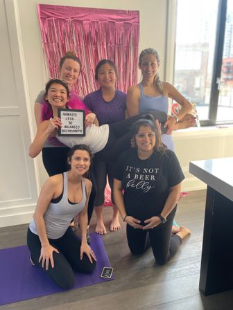 Group of six women in activewear smiling during a bachelorette yoga party in a bright urban apartment, one friend held horizontally in front of a pink foil fringe backdrop, yoga mats on a wood floor.