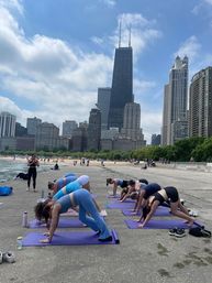 Group outdoor yoga on purple mats at the Chicago lakefront, participants in downward-dog poses with the John Hancock Center and city skyline under a partly cloudy sky