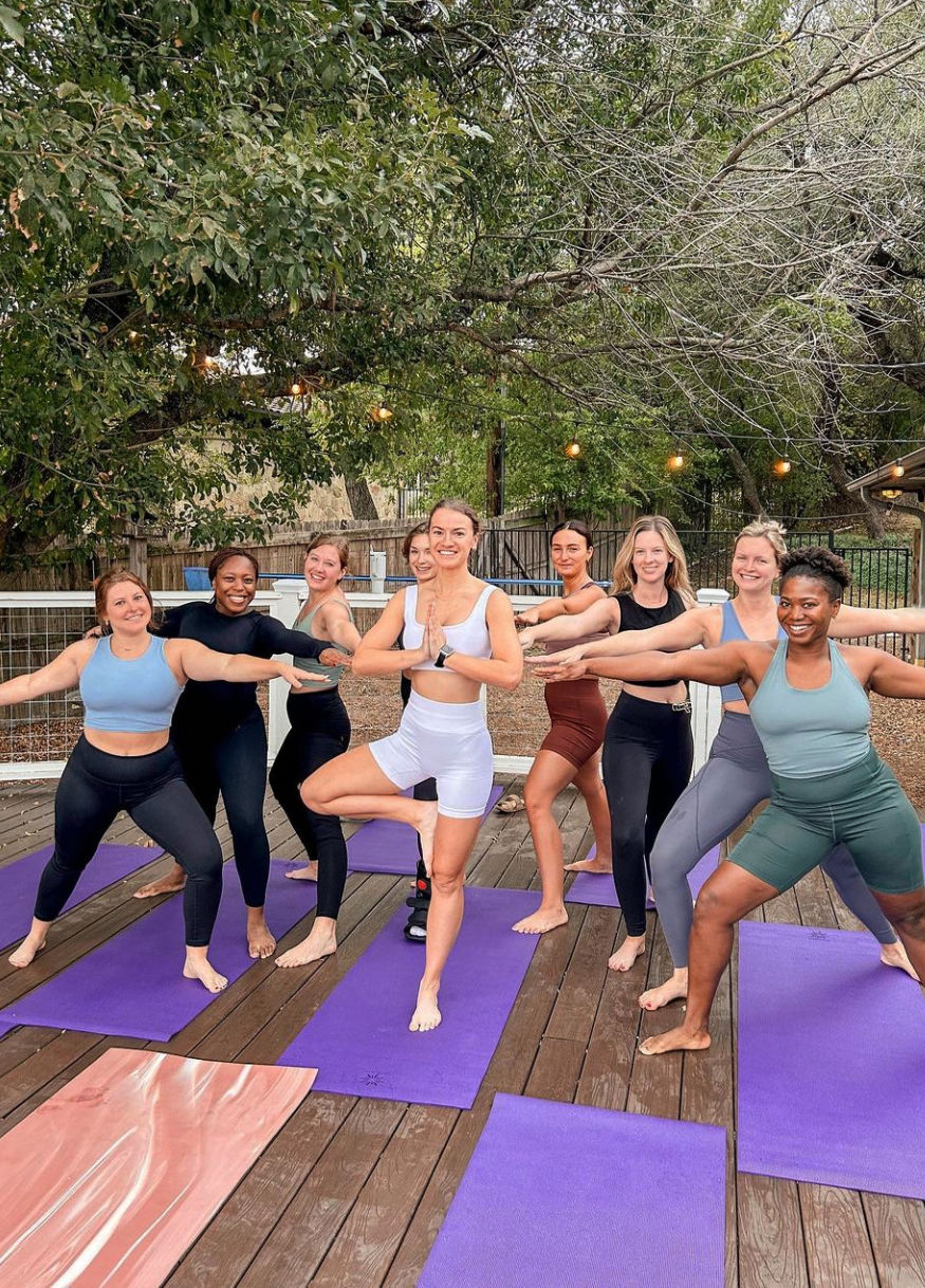 Outdoor group yoga class on a wooden deck under leafy trees — nine people in colorful athletic wear on purple mats practicing tree and warrior poses beneath string lights, community outdoor fitness scene.