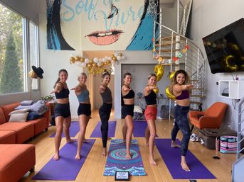 Six smiling women in athletic wear practicing a yoga warrior pose on purple mats in a bright urban loft studio with a spiral staircase, mural wall art and gold birthday balloons.