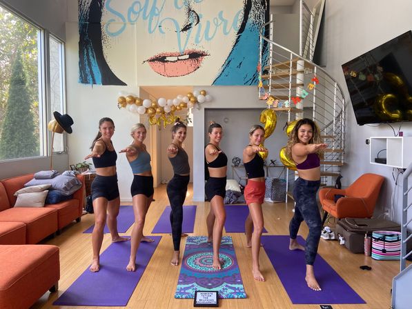 Six smiling women in athletic wear practicing a yoga warrior pose on purple mats in a bright urban loft studio with a spiral staircase, mural wall art and gold birthday balloons.