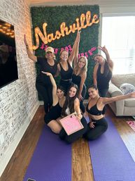 Seven women in black workout clothes smiling and posing on purple yoga mats in a bright living room, in front of a faux greenery wall with a lit “Nashville” marquee sign and holding a pink message board.