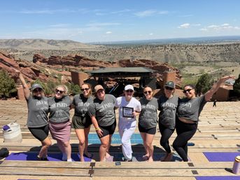 Smiling bachelorette group in matching shirts posing on wooden amphitheater seats with red sandstone formations, rolling hills and the distant Denver skyline under a bright blue sky.