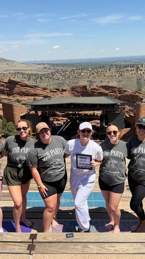 Smiling bachelorette group in matching shirts posing on wooden amphitheater seats with red sandstone formations, rolling hills and the distant Denver skyline under a bright blue sky.