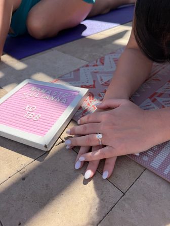 Close-up of hands with a sparkling engagement ring and white nails resting on a patterned yoga mat next to a pink letter board reading 'Namaste' on a sunlit stone patio.