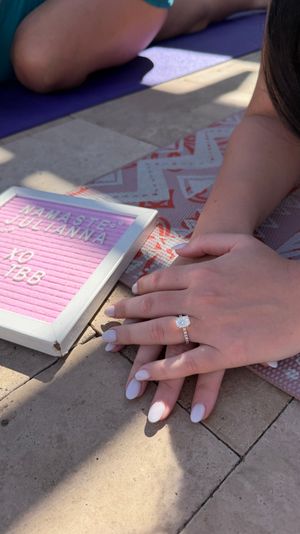 Close-up of hands with a sparkling engagement ring and white nails resting on a patterned yoga mat next to a pink letter board reading 'Namaste' on a sunlit stone patio.