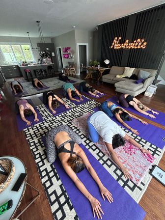 Group indoor yoga session in a cozy Nashville living-room studio — participants on purple mats in child’s pose on a patterned rug and hardwood floor beneath a lit “Nashville” sign.