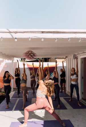 Sunny outdoor covered-patio yoga class with women in athletic wear stretching upward on purple mats in front of a pastel mural and hanging pendant light