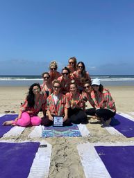 Ten women in matching watermelon-print shirts on yoga mats posing on a sunny sandy beach with ocean waves, smiling around a small letterboard at a bachelorette beach yoga gathering.