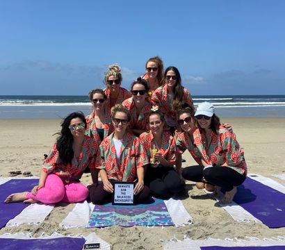 Ten women in matching watermelon-print shirts on yoga mats posing on a sunny sandy beach with ocean waves, smiling around a small letterboard at a bachelorette beach yoga gathering.
