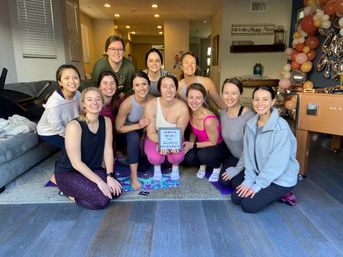 Cheerful group of women in workout clothes posing on yoga mats in a living room for a bachelorette yoga gathering, one holding a lightbox sign with balloon garland and party decor in the background.
