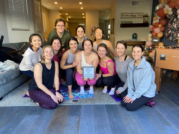 Cheerful group of women in workout clothes posing on yoga mats in a living room for a bachelorette yoga gathering, one holding a lightbox sign with balloon garland and party decor in the background.