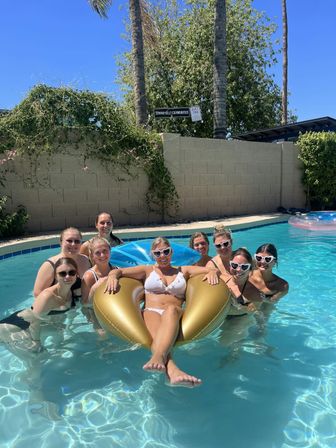 Nine friends in swimsuits posing in a sunny backyard pool, one lounging on a gold inflatable float while others surround her; clear blue sky, palm trees and a block wall in the background.
