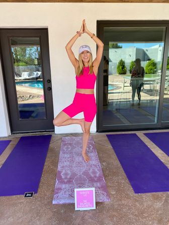 Woman in hot-pink workout set doing tree pose on a patterned yoga mat on a sunny poolside patio with purple mats and glass doors reflecting greenery