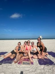 Five friends in swimsuits sitting on yoga mats on a sunny sandy beach by the ocean, smiling under a bright blue sky with a distant lifeguard tower — playful bachelorette group photo.