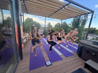Group of people doing rooftop yoga at sunset — balancing on one leg on purple mats under a wooden pergola with string lights, overlooking urban treetops.