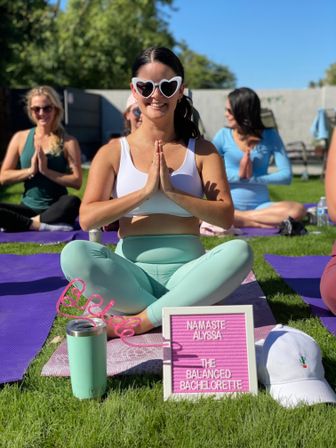 Smiling woman in heart-shaped sunglasses and mint leggings sits cross-legged in prayer pose at an outdoor lawn yoga class with purple mats, a pink “Namaste” letter board, mint tumbler and white cap nearby — playful group bachelorette wellness session