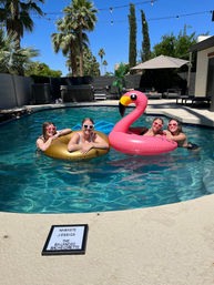 Four friends in heart-shaped sunglasses lounging on a gold inner tube and a giant pink flamingo float in a sunny backyard pool with palm trees, string lights, lounge chairs and a small letterboard on the deck.