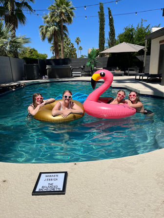 Four friends in heart-shaped sunglasses lounging on a gold inner tube and a giant pink flamingo float in a sunny backyard pool with palm trees, string lights, lounge chairs and a small letterboard on the deck.