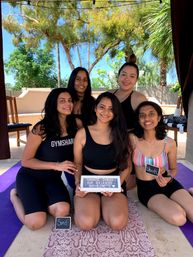 Five women in activewear smiling on yoga mats in a sunny backyard patio with palm trees and string lights, posing for a bachelorette-themed outdoor yoga celebration while holding a small sign.