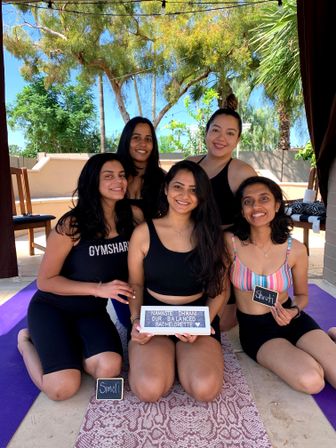 Five women in activewear smiling on yoga mats in a sunny backyard patio with palm trees and string lights, posing for a bachelorette-themed outdoor yoga celebration while holding a small sign.