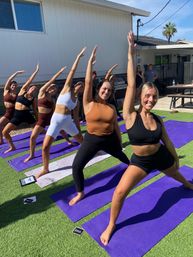 Smiling group of women practicing outdoor yoga on purple mats on artificial turf in a sunny backyard, holding side‑stretch/warrior poses