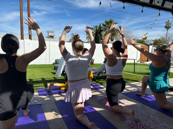 Sunny backyard yoga session with women kneeling on purple mats and raising their arms, palm trees and white block wall in the background under string lights