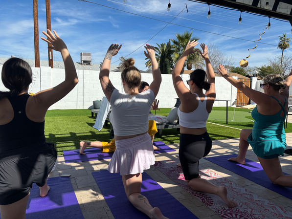 Sunny backyard yoga session with women kneeling on purple mats and raising their arms, palm trees and white block wall in the background under string lights