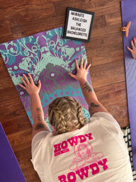 Overhead view of a person in a child's-pose on a teal-and-purple patterned yoga mat on a hardwood floor, braided hair and arm tattoos visible, wearing a bright 'Howdy Rowdy' tee with a nearby purple mat and a small letterboard displaying a bachelorette-themed message.