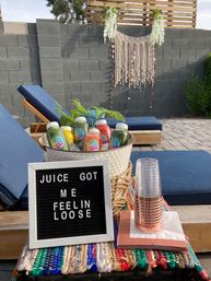 Backyard patio scene with bottled cold-pressed juices in an ice bucket, a letter board reading “JUICE GOT ME FEELIN LOOSE,” stacked cups, blue lounge chairs and a macramé wall hanging