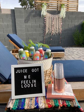 Backyard patio scene with bottled cold-pressed juices in an ice bucket, a letter board reading “JUICE GOT ME FEELIN LOOSE,” stacked cups, blue lounge chairs and a macramé wall hanging