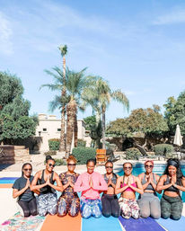 Eight women kneeling on colorful yoga mats in prayer pose by a sunny backyard pool, smiling in activewear with palm trees and desert landscaping under a clear blue sky.