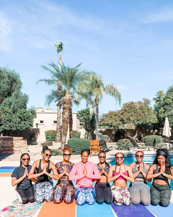 Eight women kneeling on colorful yoga mats in prayer pose by a sunny backyard pool, smiling in activewear with palm trees and desert landscaping under a clear blue sky.