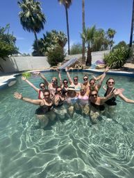 Group of women enjoying a sunny backyard pool party, smiling and waving in clear water with palm trees, a stone waterfall/slide and colorful floats under a bright blue sky.