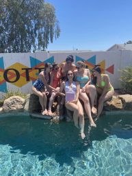 Six friends in colorful swimsuits laughing at a sunny backyard pool party, sitting on a rock ledge with feet in the water in front of a bright geometric mural on a cinderblock wall.