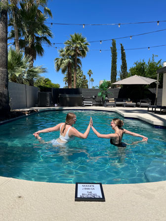 Two friends in swimsuits balancing and high‑fiving in a sunny backyard pool, surrounded by palm trees, string lights, umbrella and poolside lounge for a playful summer scene.