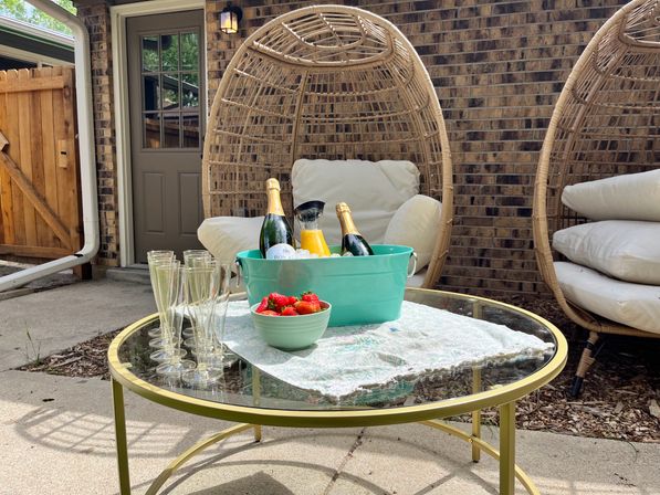 Cozy brick-walled backyard patio with rattan egg chairs and a glass-top gold table set for mimosas — teal tub of chilled sparkling wine and orange juice, bowl of strawberries, and champagne flutes.