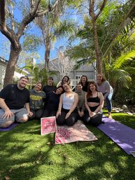 Smiling group of people seated on yoga mats in a sunny backyard garden with palm trees, posing around a small congratulatory bachelorette sign.