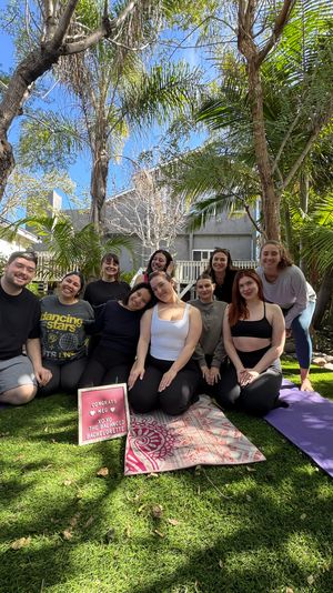 Smiling group of people seated on yoga mats in a sunny backyard garden with palm trees, posing around a small congratulatory bachelorette sign.
