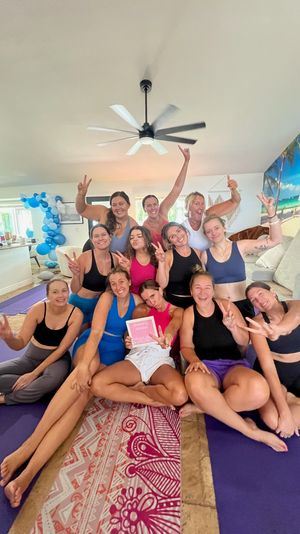 Happy group of women on purple yoga mats posing with peace signs in a bright living-room yoga retreat, tropical beach wall mural and blue balloon arch in the background