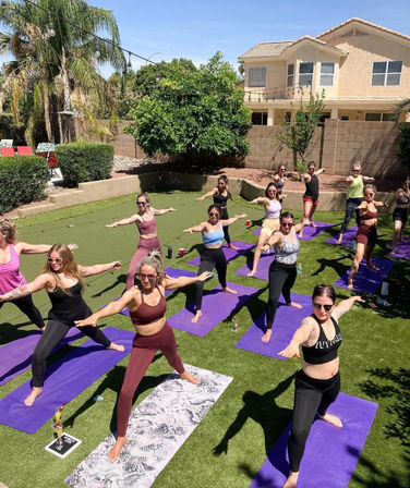 Group outdoor yoga class on purple mats in a sunny suburban backyard, women practicing Warrior pose near palm trees and a stucco house.