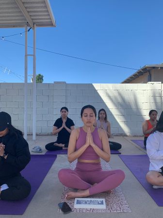 Sunny outdoor group yoga class under a clear blue sky — seated meditation with a woman in mauve activewear at center, others on purple mats in front of a white cinderblock wall