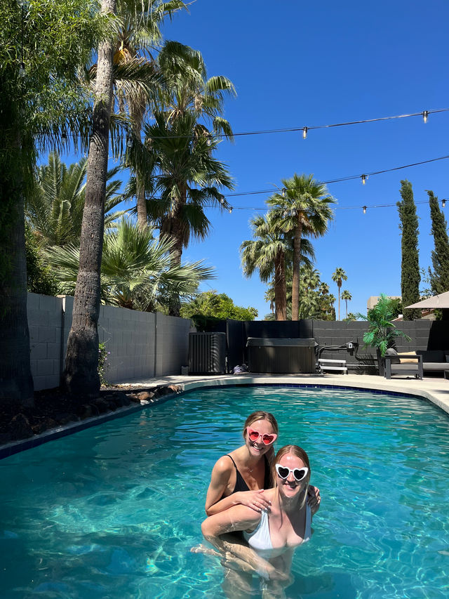 Two friends wearing heart-shaped sunglasses laughing in a sunny backyard pool framed by palm trees and string lights